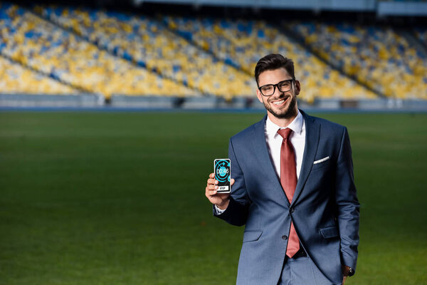 smiling young businessman in suit and glasses holding smartphone with stopwatch app at stadium