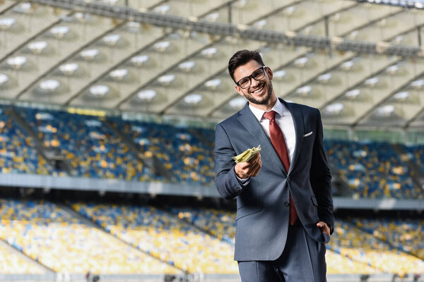 smiling young businessman in suit and glasses giving money at stadium