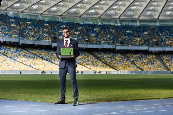 Takım elbiseli gülümseyen genç işadamı stadyumdaki futbol formasyonunda dizüstü bilgisayarı gösteriyor.