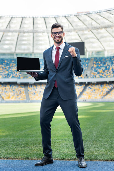 smiling young businessman in suit showing yeah gesture while holding laptop with blank screen at stadium