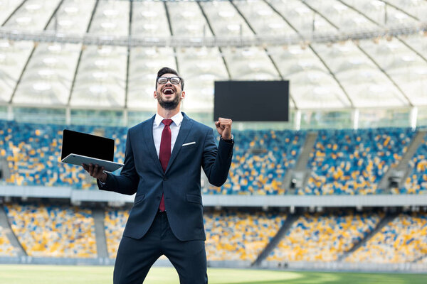 низкий угол обзора счастливого молодого бизнесмена в костюме, показывающего жест "да" и держащего ноутбук с пустым экраном на стадионе
