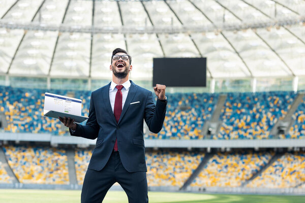 KYIV, UKRAINE - JUNE 20, 2019: low angle view of happy young businessman in suit showing yes gesture and holding laptop with facebook website at stadium