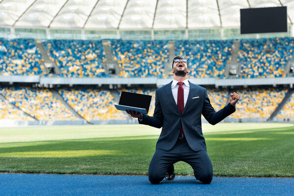 happy young businessman in suit showing yes gesture and holding laptop with blank screen while standing on knees at stadium