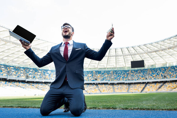 счастливый молодой бизнесмен в костюме держит ноутбук с чистым экраном и деньгами, стоя на коленях на стадионе
