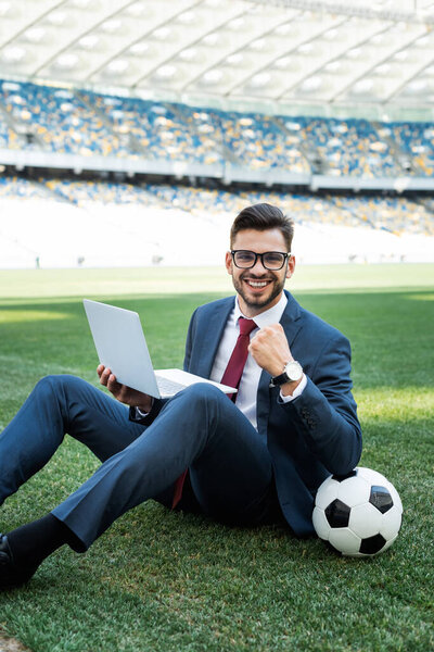 smiling young businessman in suit with laptop and soccer ball sitting on football pitch and showing yes gesture at stadium, sports betting concept