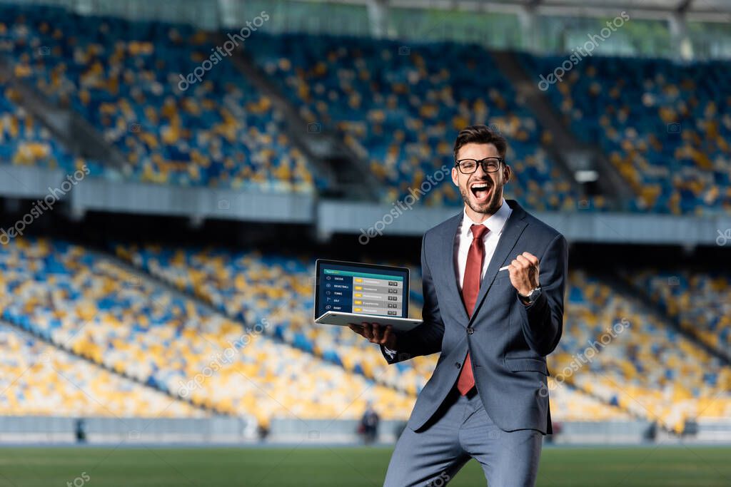Happy young businessman in suit holding laptop with sport betting website and showing yes gesture at stadium