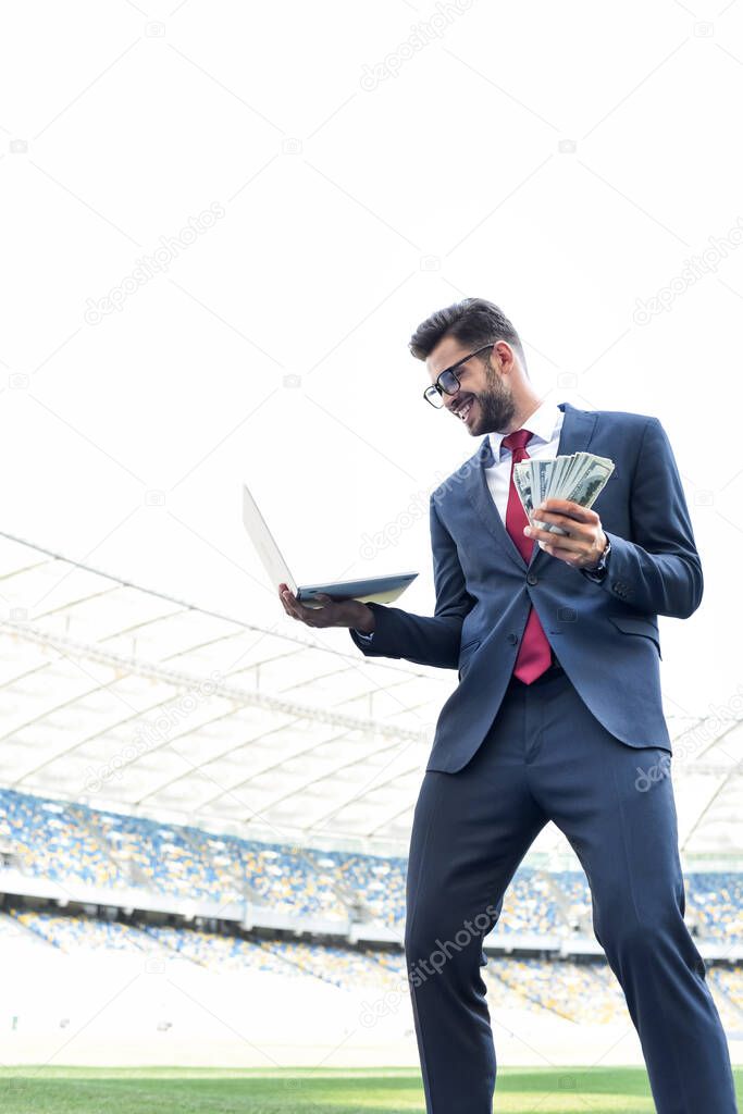 low angle view of happy young businessman in suit holding laptop and money at stadium, sports betting concept