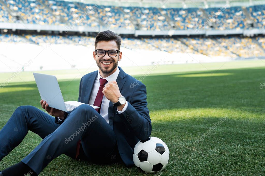 smiling young businessman in suit with laptop and soccer ball sitting on football pitch and showing yes gesture at stadium, sports betting concept