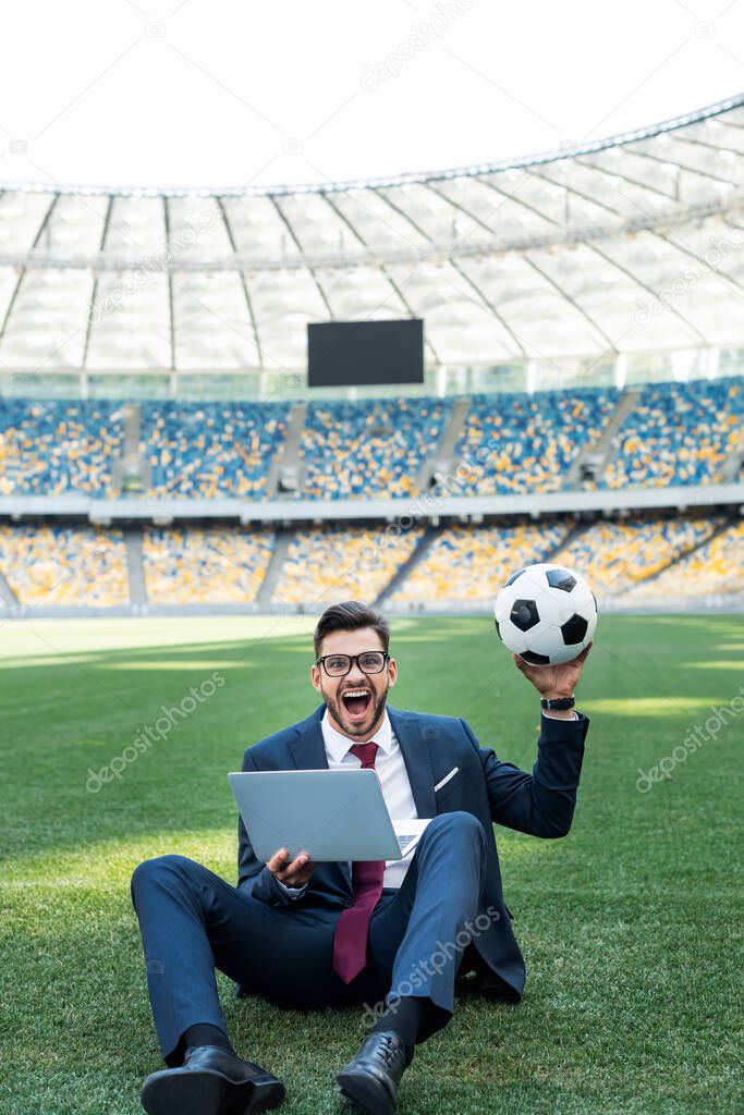 happy young businessman in suit with laptop and soccer ball sitting on football pitch at stadium, sports betting concept