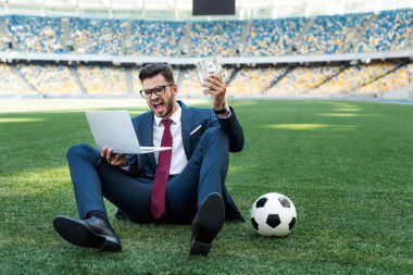 Takım elbiseli genç işadamı, dizüstü bilgisayarı, parası ve futbol topuyla stadyumdaki futbol sahasında oturuyor ve bağırıyor, spor bahisleri oynuyor.