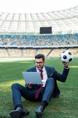 Takım elbiseli genç iş adamı dizüstü bilgisayar ve futbol topuyla stadyumdaki futbol sahasında oturuyor ve bağırıyor, spor bahisleri oynuyor.