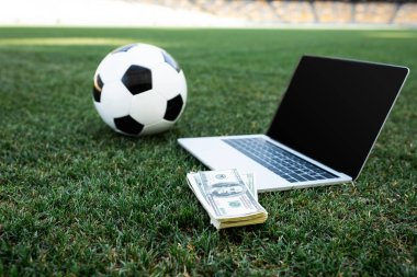 Stadyumdaki çimenli futbol sahası üzerinde boş ekranı olan futbol topu, para ve dizüstü bilgisayarın seçici odak noktası, online bahis kavramı