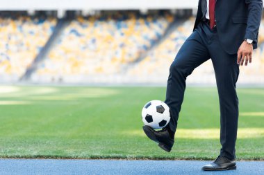cropped view of young businessman in suit playing with soccer ball at stadium clipart