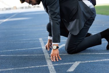 cropped view of young businessman in suit in start position on running track at stadium clipart