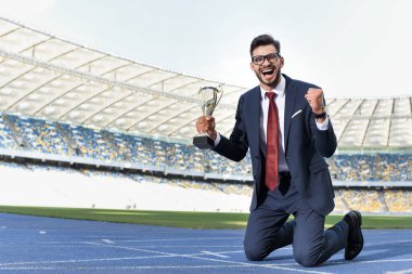 happy young businessman in suit standing on knees on running track with trophy at stadium clipart