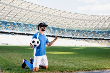Profesyonel futbolcu, VR kulaklıklı, mavi-beyaz üniformalı, top dizlerinin üstünde duruyor ve stadyumdaki futbol sahasına evet işareti yapıyor.