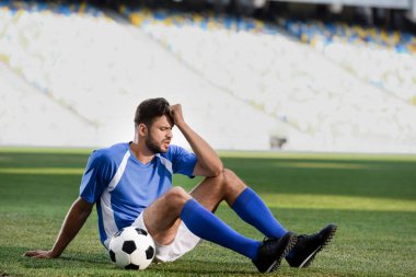 sad professional soccer player in blue and white uniform sitting with ball on football pitch at stadium clipart