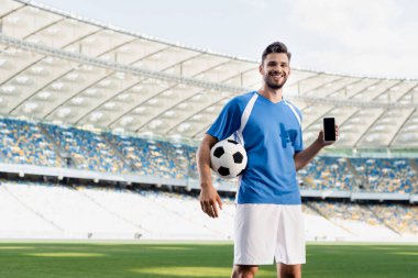 Mavi ve beyaz üniformalı gülümseyen profesyonel futbolcu stadyumdaki boş ekranlı akıllı telefonu gösteriyor.