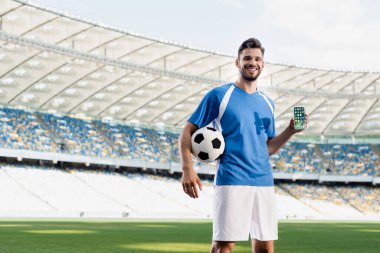 KYIV, UKRAINE - 20 Haziran 2019: Stadyumdaki iPhone uygulamalarıyla akıllı telefonu gösteren mavi ve beyaz üniformalı gülümseyen profesyonel futbolcu