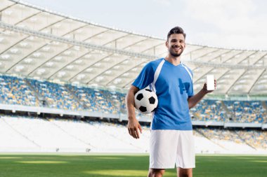 Mavi ve beyaz üniformalı gülümseyen profesyonel futbolcu stadyumdaki boş ekranlı akıllı telefonu gösteriyor.