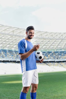 Stadyumda akıllı telefon kullanan mavi ve beyaz üniformalı gülümseyen profesyonel futbolcu.