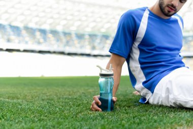 Futbol sahasında oturan mavi beyaz üniformalı profesyonel futbolcunun stadyumdaki spor şişesi manzarası.