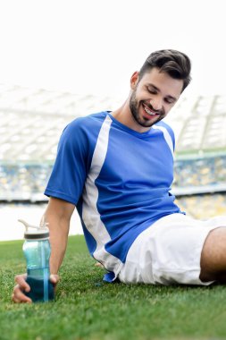 Mavi ve beyaz formalı gülümseyen profesyonel futbolcu stadyumdaki spor şişesiyle futbol sahasında oturuyor.