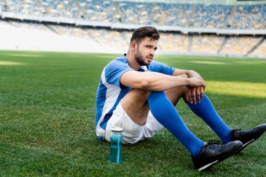 Futbol sahasında mavi ve beyaz formalı profesyonel futbolcu stadyumda spor şişesiyle oturuyor.