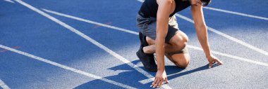 partial view of handsome runner in start position on running track at stadium, panoramic shot clipart