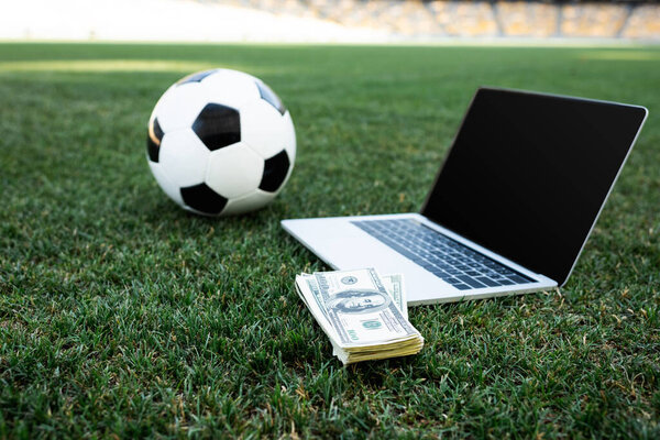 selective focus of soccer ball, money and laptop with blank screen on grassy football pitch at stadium, online betting concept