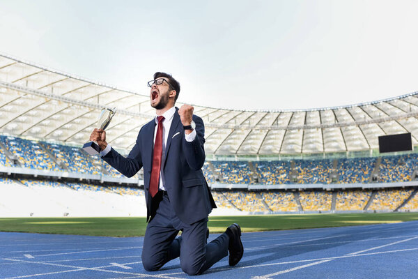young businessman in suit standing on knees on running track with trophy and scream at stadium