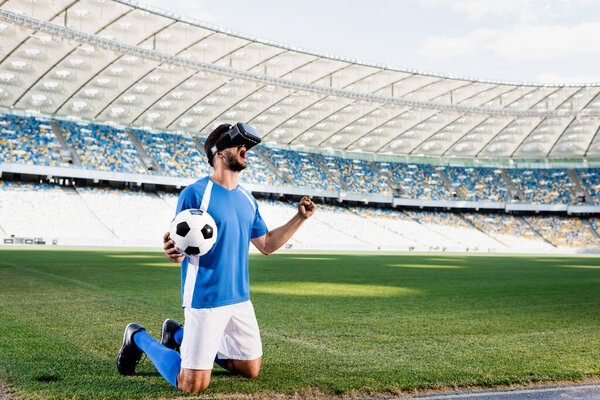 professional soccer player in vr headset and blue and white uniform with ball standing on knees and showing yes gesture on football pitch at stadium