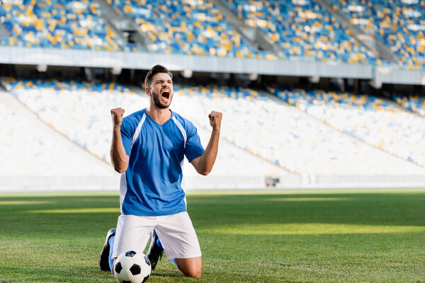 emotional professional soccer player in blue and white uniform with ball standing on knees on football pitch and showing yes gesture at stadium