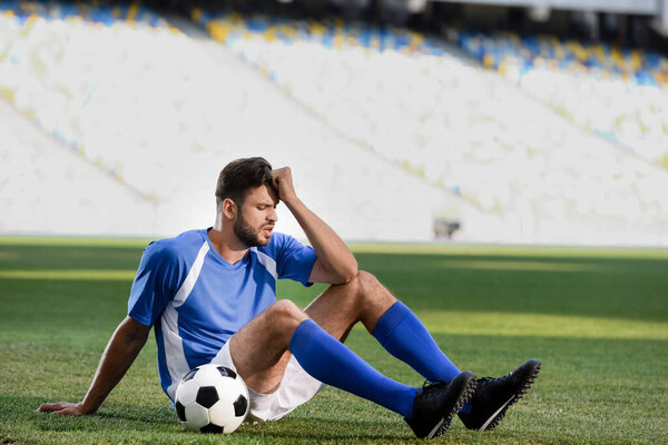 sad professional soccer player in blue and white uniform sitting with ball on football pitch at stadium