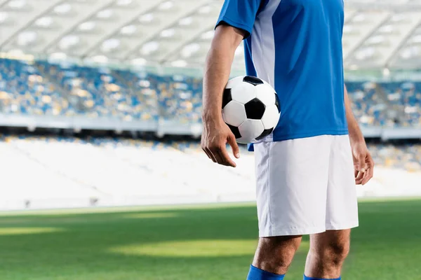 cropped view of professional soccer player in blue and white uniform with ball on football pitch at stadium