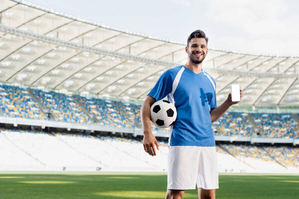 smiling professional soccer player in blue and white uniform with ball showing smartphone with blank screen at stadium