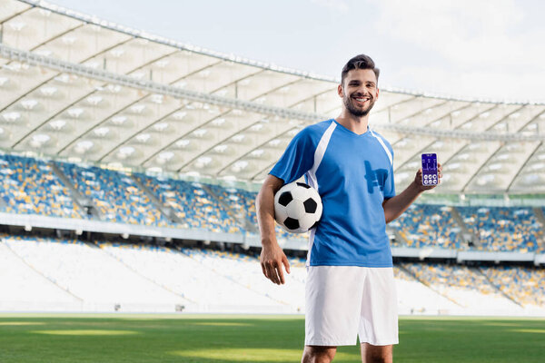 professional soccer player in blue and white uniform with ball showing smartphone with heartbeat rate on screen at stadium