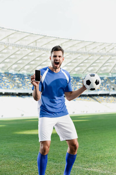 professional soccer player in blue and white uniform with ball showing smartphone with blank screen and shouting at stadium