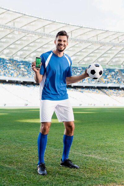 smiling professional soccer player in blue and white uniform with ball showing smartphone with heartbeat rate on screen  at stadium