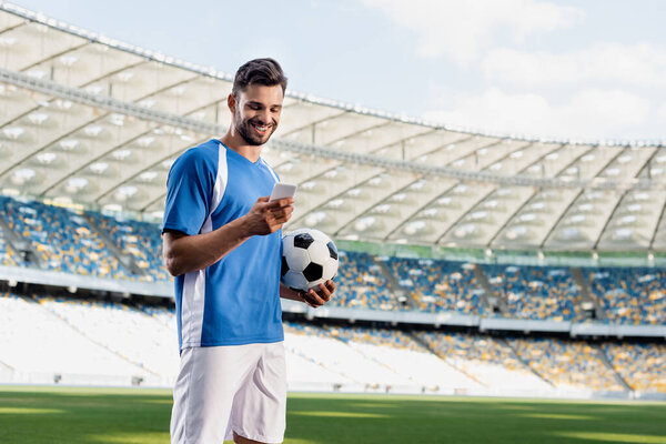 smiling professional soccer player in blue and white uniform with ball using smartphone at stadium