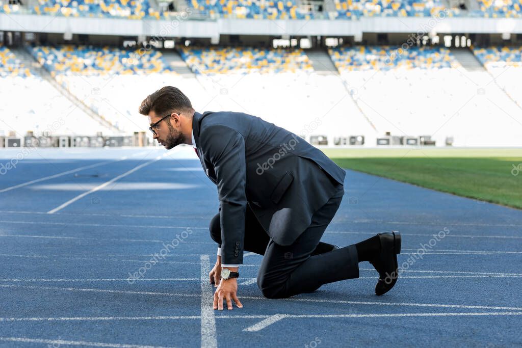 side view of young businessman in suit in start position on running track at stadium