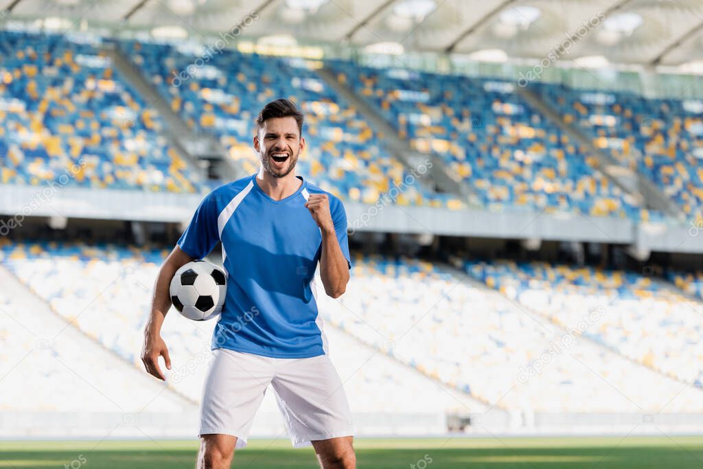 happy professional soccer player in blue and white uniform with ball showing yes gesture on football pitch at stadium