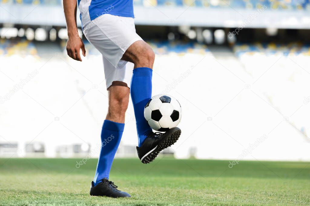 cropped view of professional soccer player in blue and white uniform playing with ball on football pitch at stadium