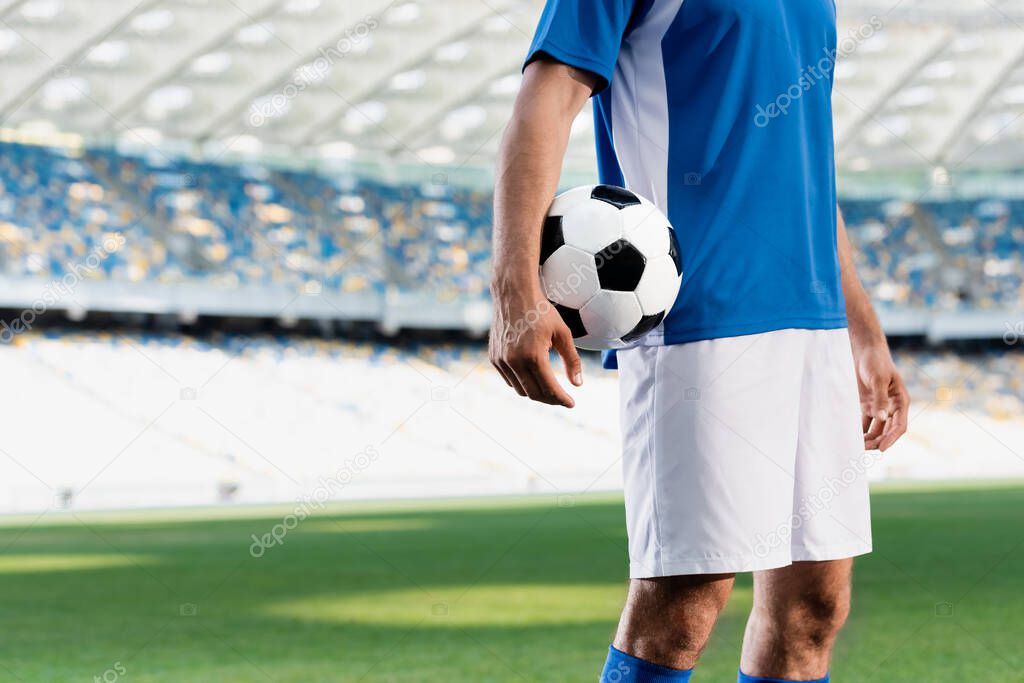 cropped view of professional soccer player in blue and white uniform with ball on football pitch at stadium