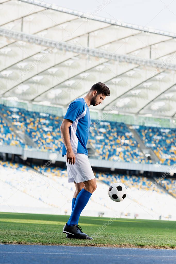 side view of professional soccer player in blue and white uniform with ball on football pitch at stadium