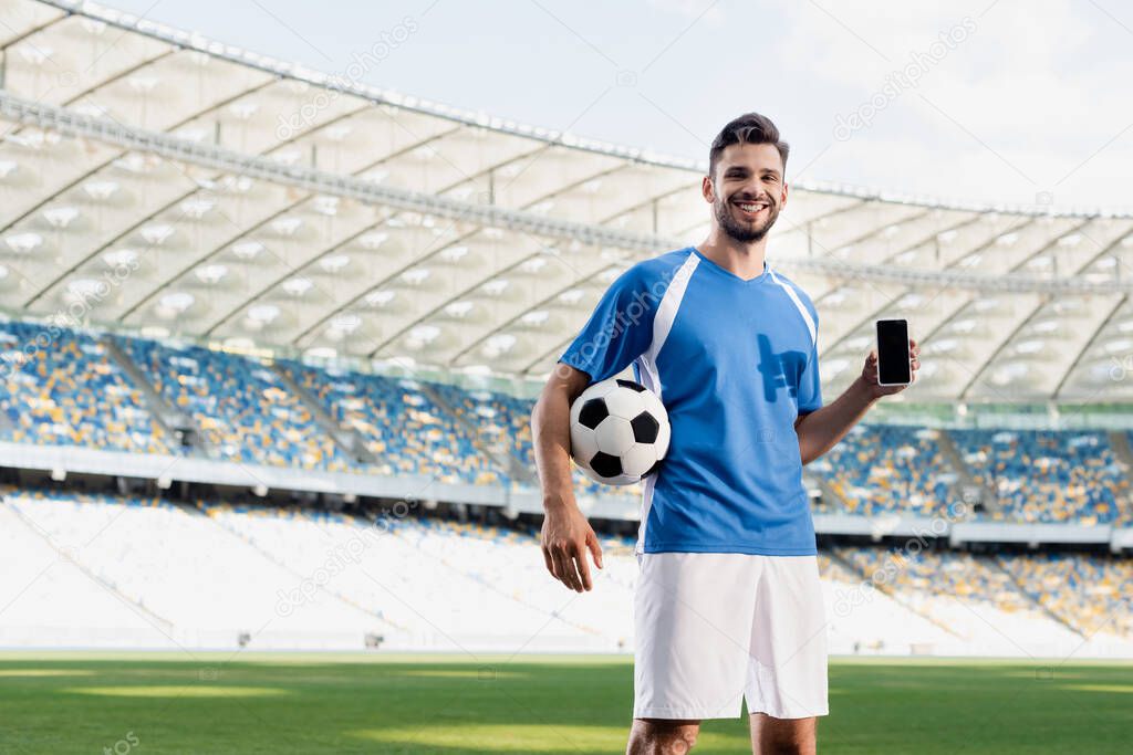 smiling professional soccer player in blue and white uniform with ball showing smartphone with blank screen at stadium