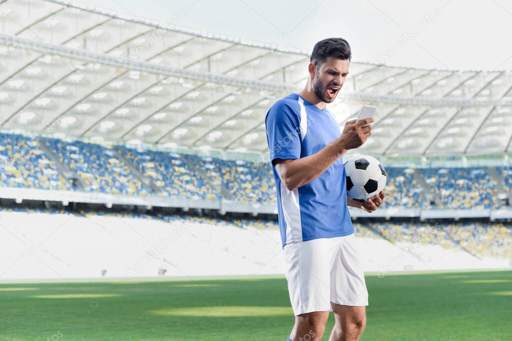 emotional professional soccer player in blue and white uniform with ball using smartphone at stadium