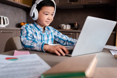 gülümseyen Asyalı çocuk evde karantina sırasında kitaplar, dizüstü bilgisayarlar ve kulaklıklarla çalışıyor.