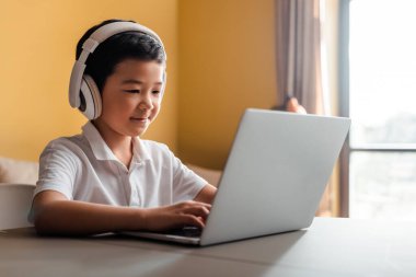 Karantina sırasında evde dizüstü bilgisayar ve kulaklıkla çalışan gülümseyen Asyalı çocuk.
