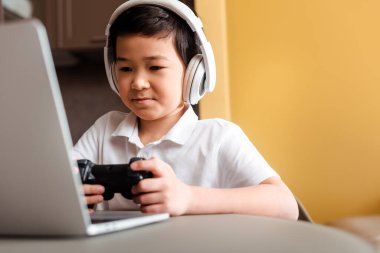 KYIV, UKRAINE - 22 Nisan 2020: joystick ve kendi kendini izole eden dizüstü bilgisayarla oyun oynayan yakışıklı Asyalı çocuk